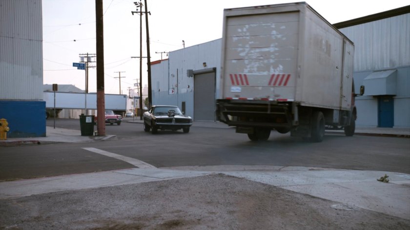 Lola chasing black Charger along street towards backing truck.