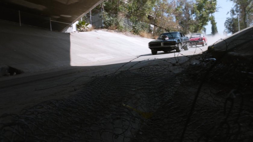 Lola chasing black Charger along river culvert.