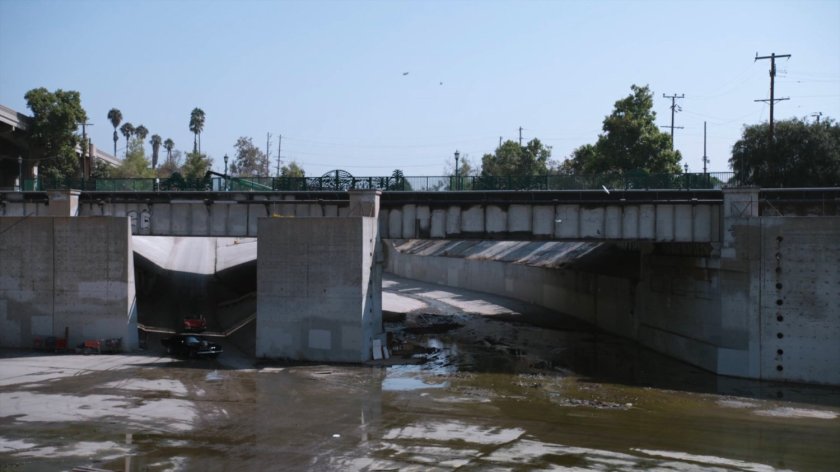 Lola chasing black Charger along river culvert.