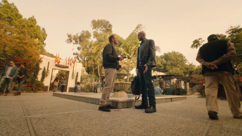 Coulson talks with Mack by fountain in Spanish plaza.