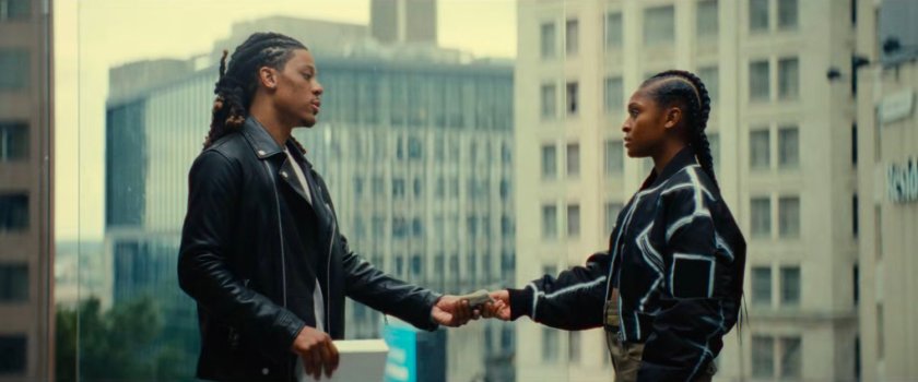 Riri taking money from a man by a large window with downtown outside.