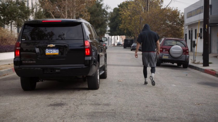 An SUV pulling up next to a figure in a hoodie jogging along a street.